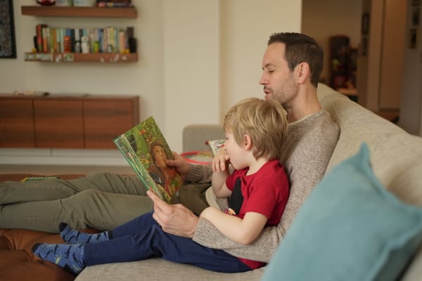 Father and boy reading a storybook on the sofa, cushions stacked around them