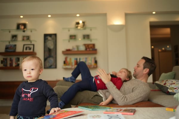 Toddler standing at the table while father and boy play on the sofa in the background