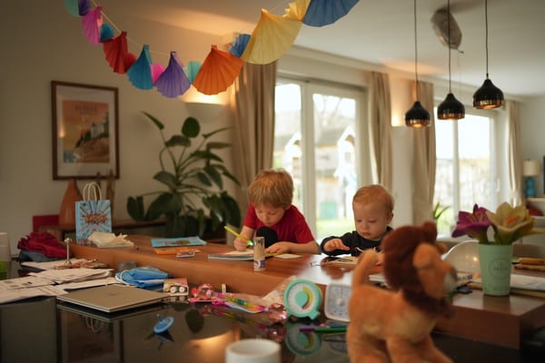Two children drawing at the dining table under colorful paper fan garlands