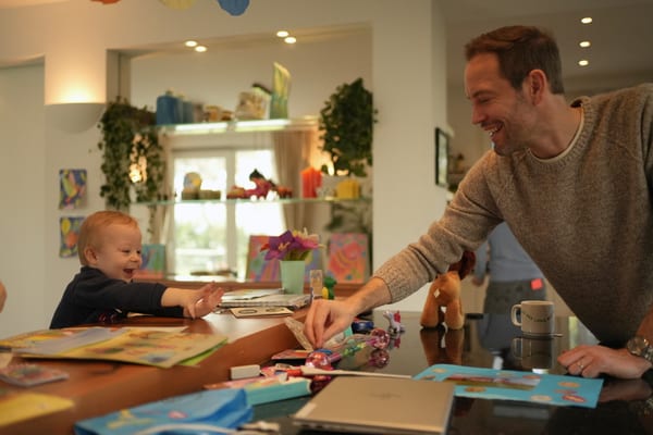 Father laughing with a toddler reaching across the craft-covered dining table