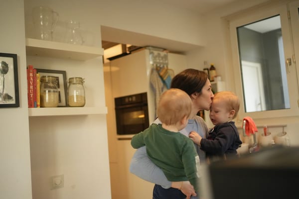 Mother kissing a toddler on the forehead while carrying another child in the kitchen