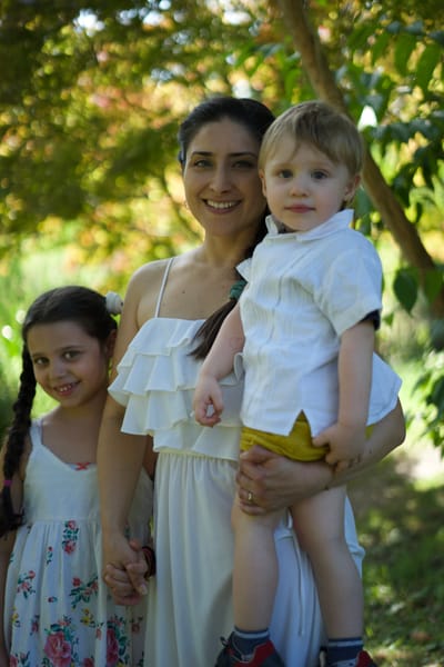 Mother holding toddler son with daughter by her side in a sunlit garden