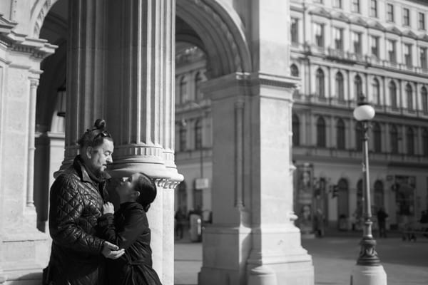 Couple embracing beside a stone column, with a street lamp and old facades behind