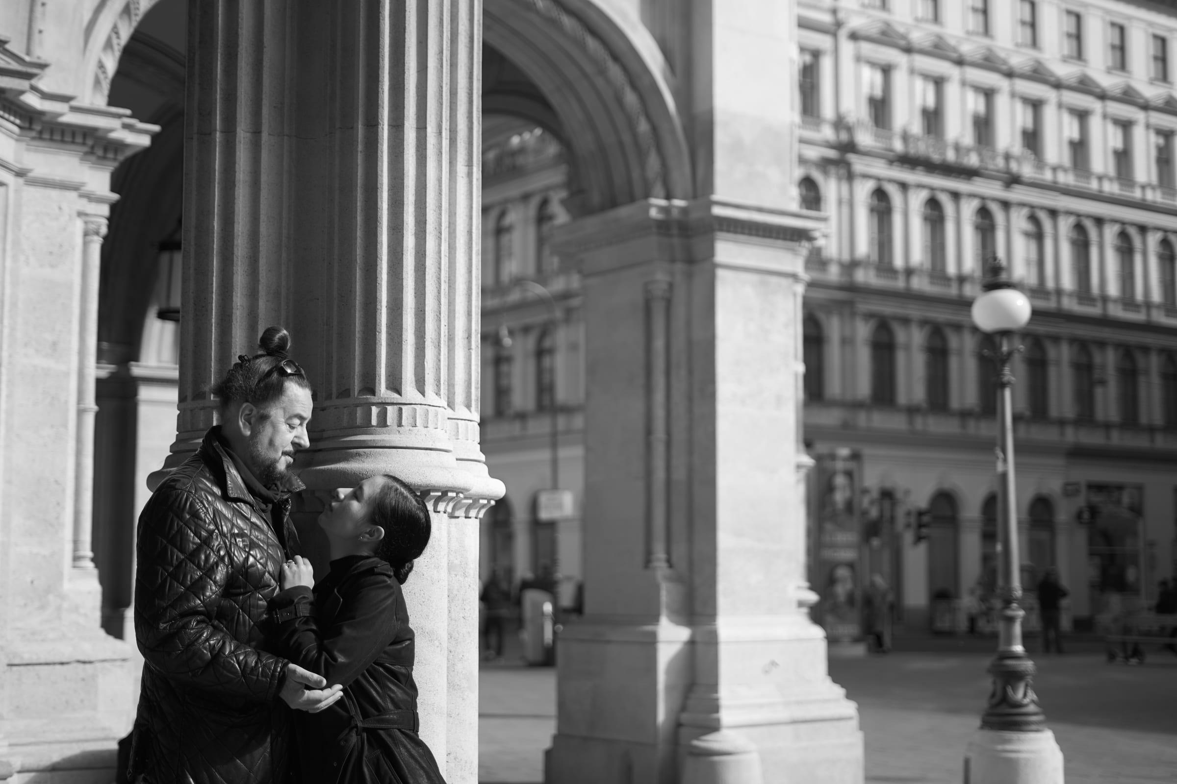 Couple embracing beside a stone column, with a street lamp and old facades behind