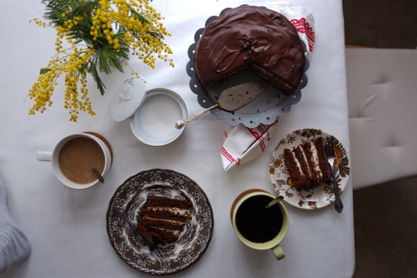 Chocolate ganache cake on a scalloped stand with coffee cups, mimosa flowers, and folk tea towel