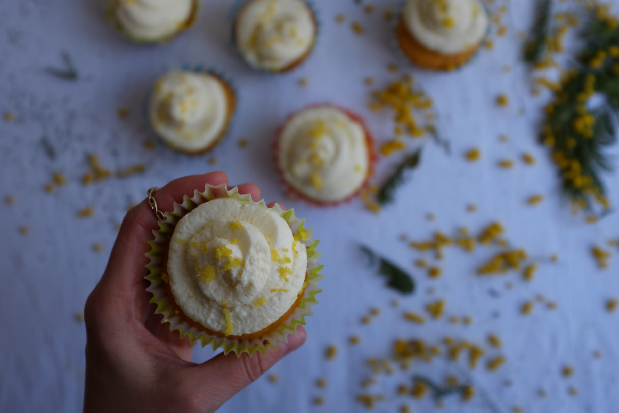 Hand holding a lemon cupcake with cream frosting and zest, mimosa flowers on blue linen behind