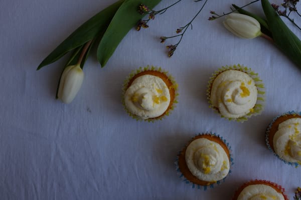 Five lemon cupcakes in pastel liners on pale linen with white tulips and lavender sprigs