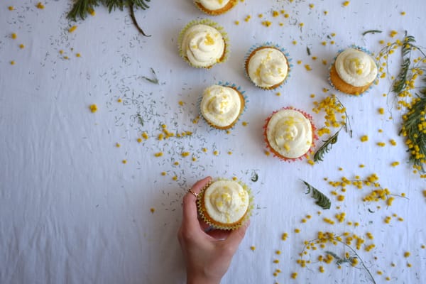 Hand reaching for a lemon cupcake among seven scattered on white linen with yellow mimosa branches