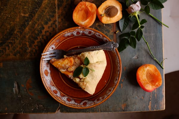Slice of apricot galette on brown ceramic plate with fresh apricots and pink rose on weathered wood
