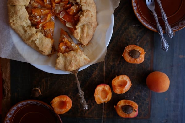 Whole apricot galette with almond topping on parchment, silver cake server, halved apricots on dark wood