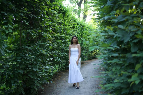 Woman in a white dress standing on a gravel path between tall green hedges