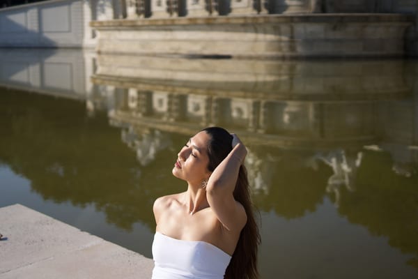 Woman with eyes closed in the sun, the fountain wall reflected in the water behind her