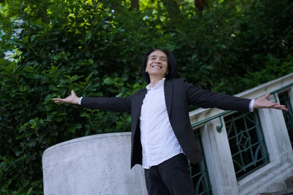 Man with arms spread wide by a stone balustrade, green hedge behind