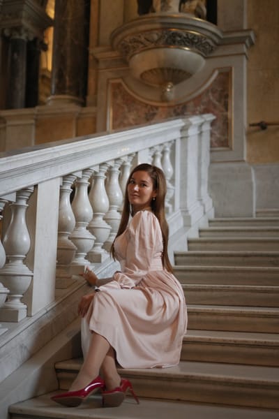 Seated portrait on marble stairs with pearl choker and red heels, warm museum lighting