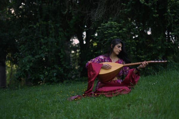 Woman in crimson and purple embroidered traditional robe sitting on park grass playing a saz