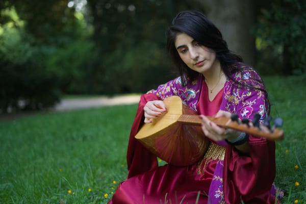 Woman sitting on park grass in embroidered traditional dress, close-up playing a saz with focused downward gaze