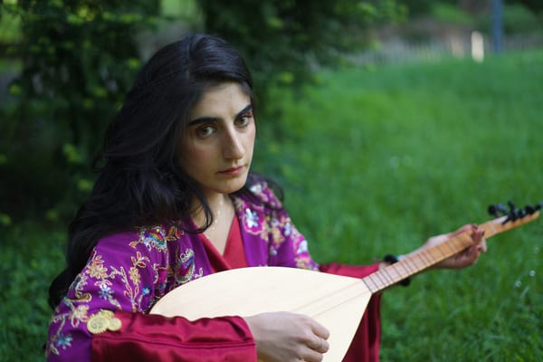 Woman in embroidered traditional dress playing a saz on park grass, direct gaze at camera