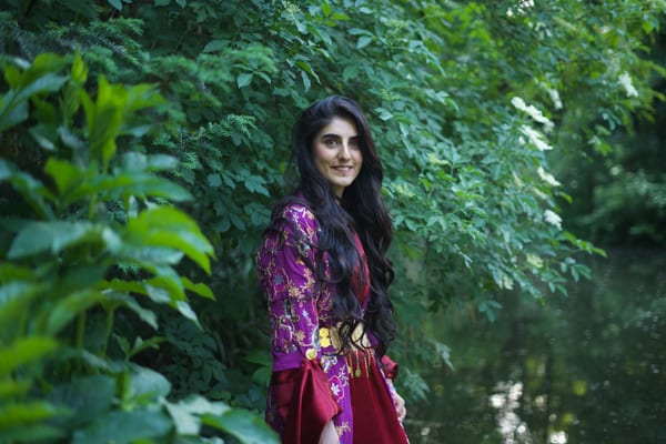 Woman in crimson traditional robe standing by a Vienna park pond, leafy branches framing the scene, smiling