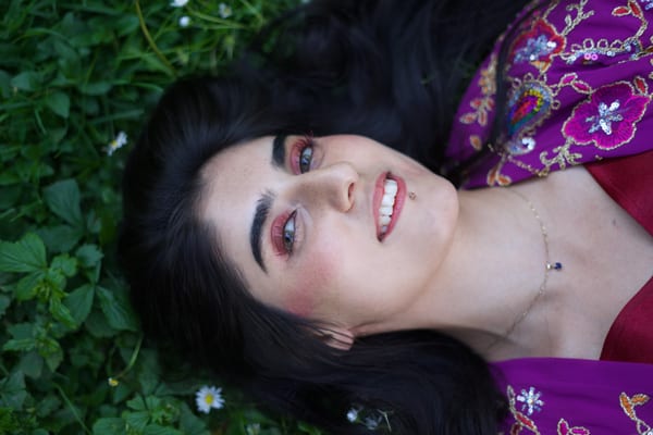 Overhead close-up of smiling woman lying among park clover and daisies, embroidered robe visible at the edges