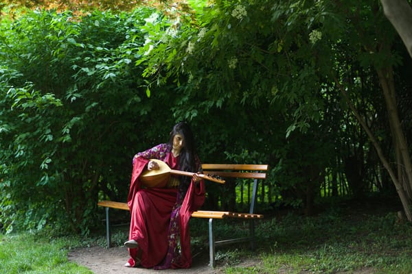 Woman in crimson robe seated on a park bench playing saz, lush green trees and dappled shade behind her