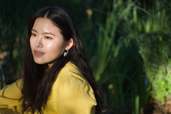 Woman in a yellow dress looking to the side, sun on her face and green leaves behind