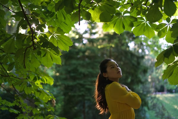 Woman in yellow looking up under sunlit chestnut leaves, hand on her chest