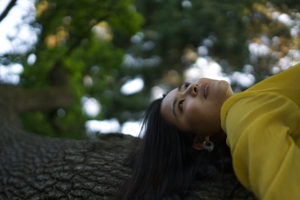 Woman in yellow lying back on a large tree trunk, looking up at the leaves