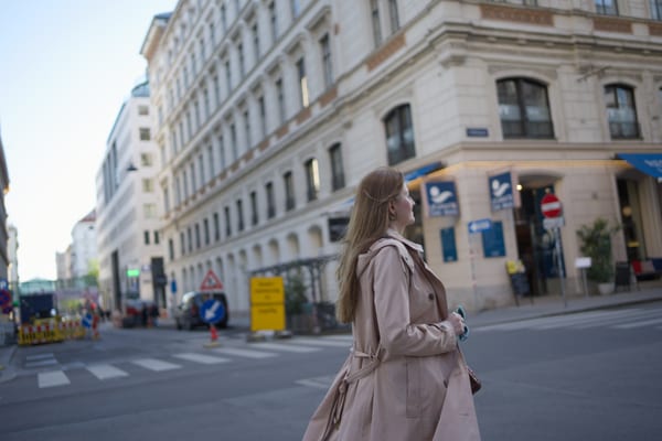 Woman in pink trench coat walking away on a Vienna street, classical building and clear blue sky behind