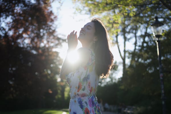 Woman in floral dress backlit by afternoon sun, hands near face, park trees behind