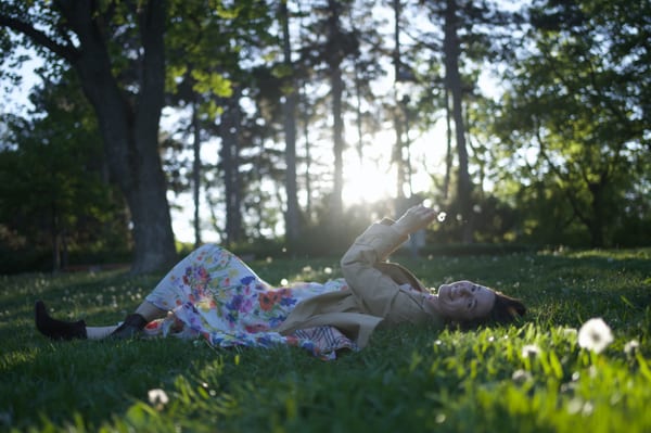 Woman lying on park grass in floral dress, sunflare through trees, relaxed pose