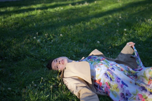Woman resting on grass in floral dress and beige jacket, smiling under dappled light