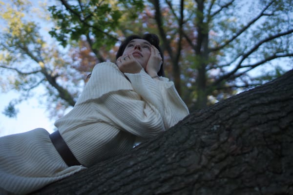 Woman on tree branch in white knit outfit, looking upward, autumn foliage behind