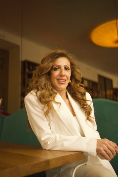 Woman in white blazer smiling at the camera, leaning on a wooden table in a warm Vienna café