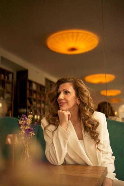 Woman in white blazer resting her chin on her hand, looking sideways in a cozy Vienna café with golden pendant lights