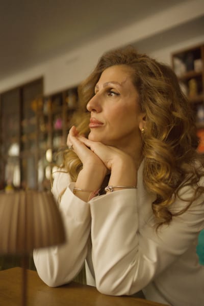 Close-up of a woman with curly hair resting her face on clasped hands, gazing upward in warm café light